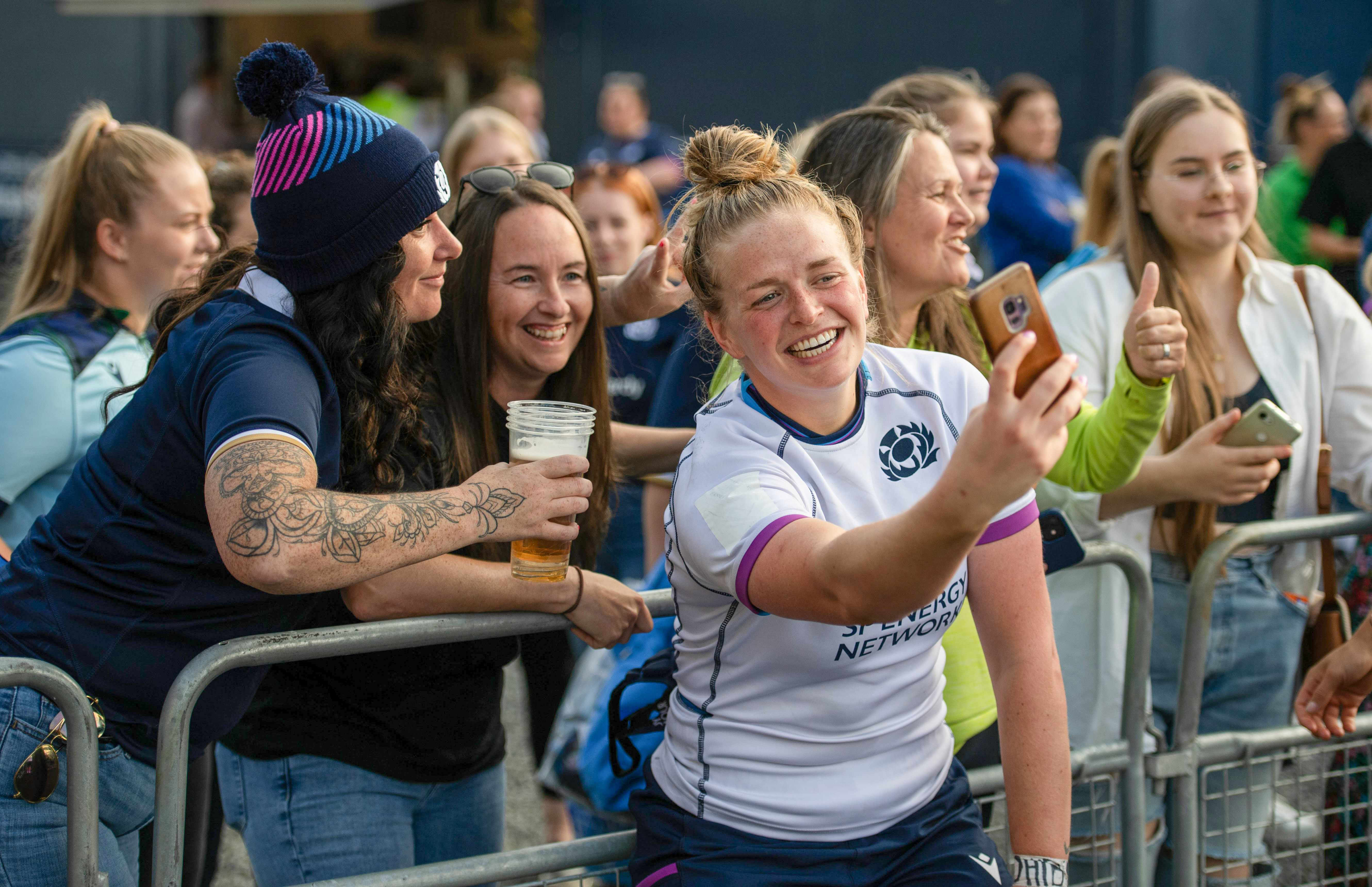 Image shows a rugby player taking a selfie on her mobile, with the crowd behind metal barriers.